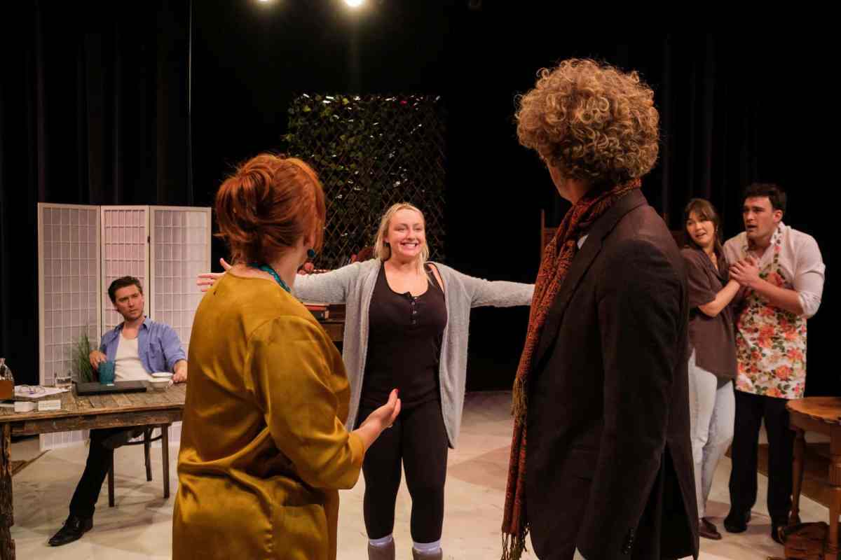 A fair-haired woman in a long grey cardigan over black clothes stands with arms outstretched on centre stage. She is framed by two actors with their backs to the camera, red-haired woman on the left and a brown-haired man on the right.