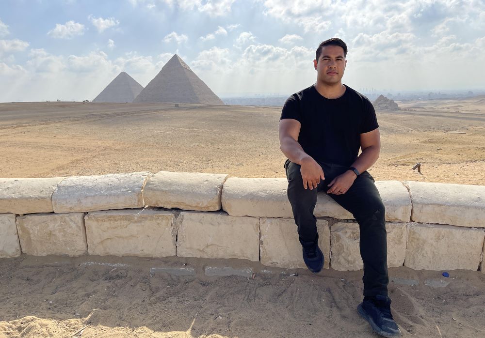 An Indian-Australian Man Dressed All In Black Sits On A Low Stone Wall. Behind Him In The Distance Stand The Pyramids, Rising Up From A Desert Landscape.