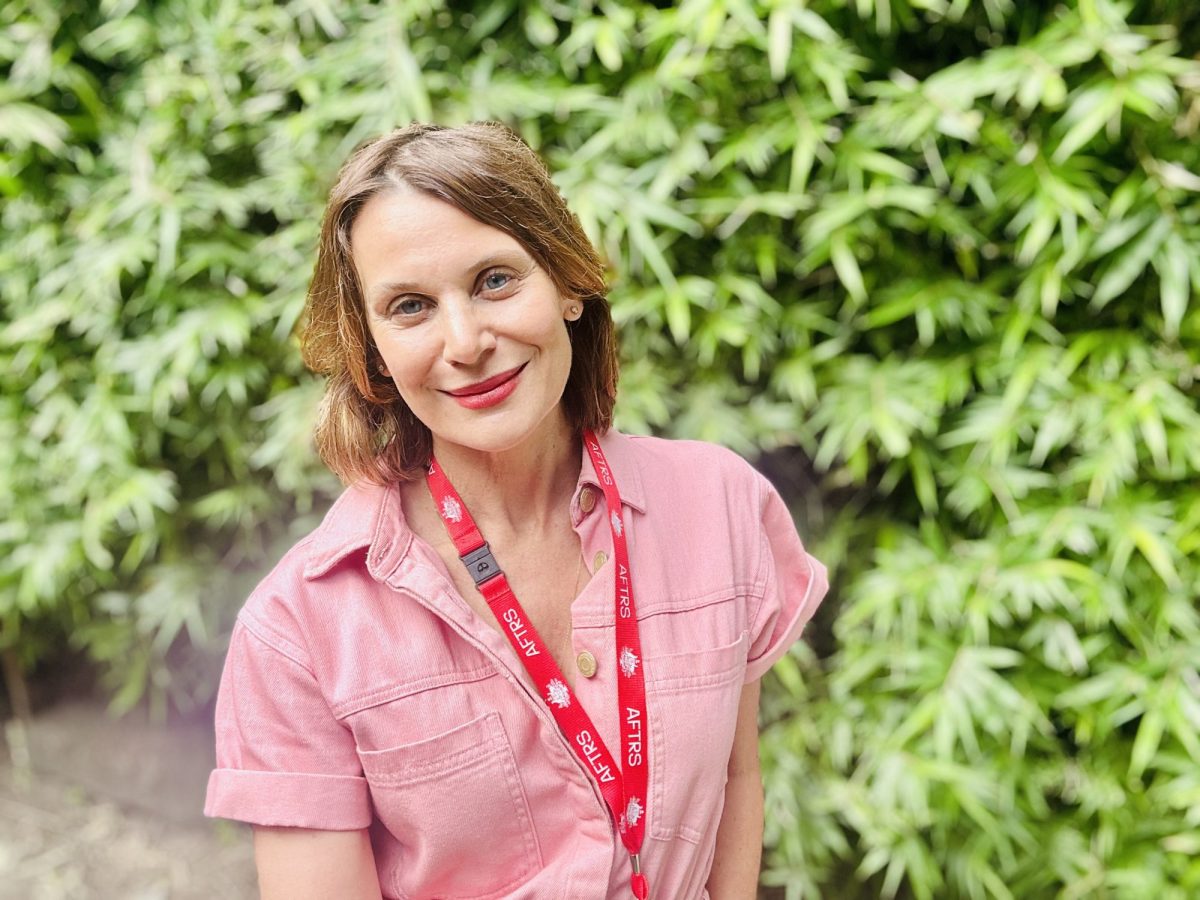 Diana Glenn. A smiling middle-aged woman with a brown crop and wearing a short sleeved pink shirt, with an AFTRS lanyard stands in front of green foliage.